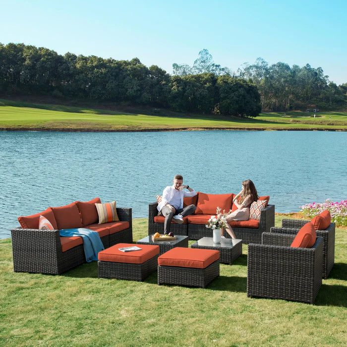 A complete outdoor patio furniture set, featuring dark brown wicker and burnt orange cushions, is arranged on a lush green lawn overlooking a tranquil lake. 